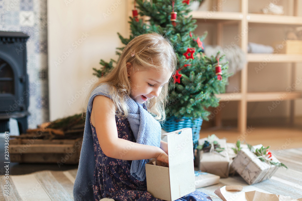 Happy child girl opens gift box under Christmas tree. Kid is surprised. Natural children emotions. Cozy warm home, living room, blue decorating. Concept of giving presents, New Year. Lifestyle moment.