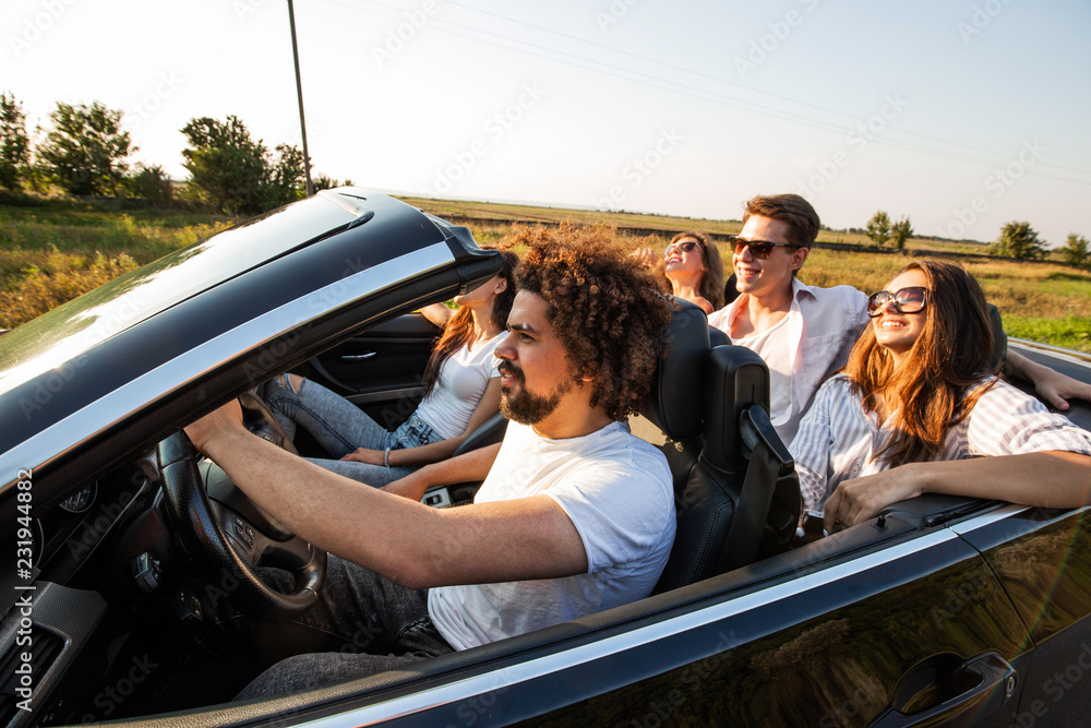 Naklejka premium Group of young beautiful girls and guys in sunglasses smile and ride in a black cabriolet on the road on a sunny day.