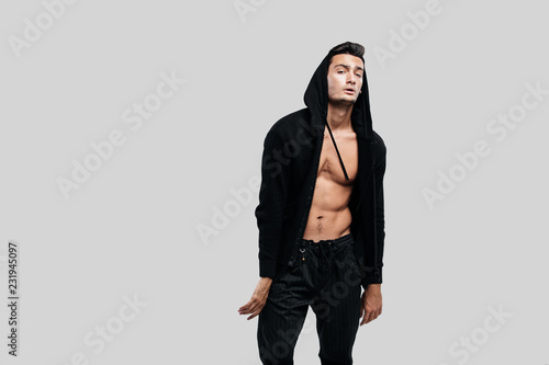 Stylish young dancer dressed in a sweatshirt on a naked torso with a hood and black pants stands on a white background