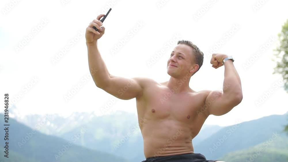 Handsome bodybuilder with perfect muscular body and blonde hair holding smart phone and taking a selfie outdoors over beautiful mountains with green flora on background. Handsome bodybuilder with perfect muscular body and blonde hair holding smart phone and taking a selfie outdoors over beautiful mountains with green flora on background.