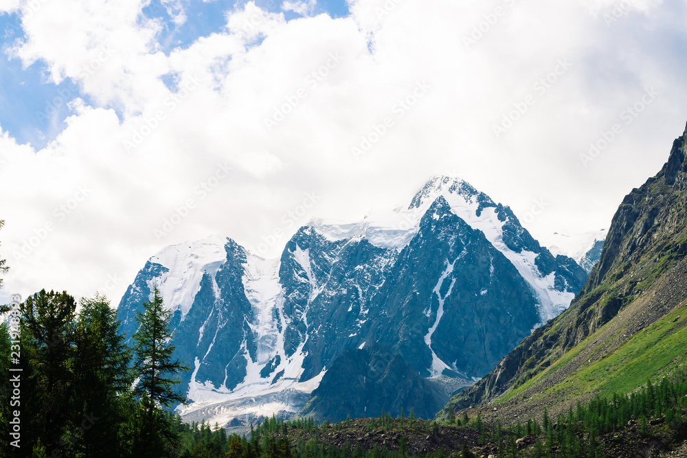 Fototapeta premium Snowy mountain top between rocky mountains under overcast sky. Rocky ridge in mist above forest. Atmospheric minimalistic landscape of majestic nature.