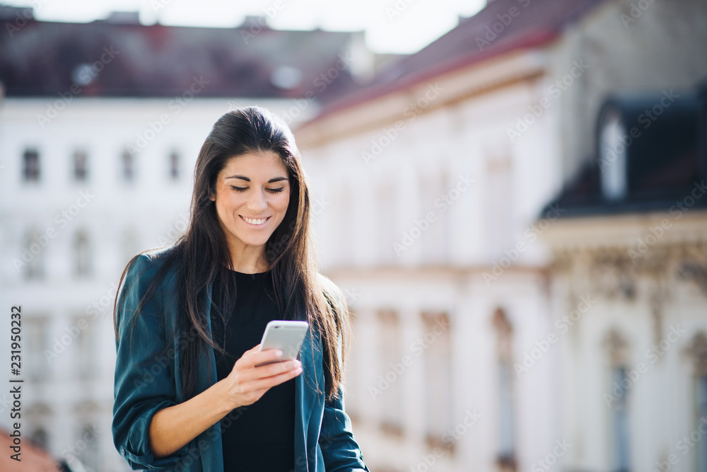 Young businesswoman with smartphone standing on a terrace outside an office in city.