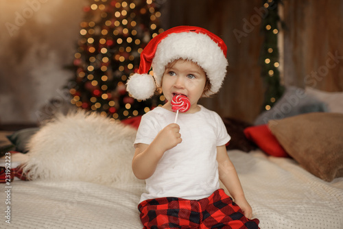 Little cute baby girl in Santa Claus hat is sitting in the room on the background of the Christmas tree and eating sweet candy on a stick