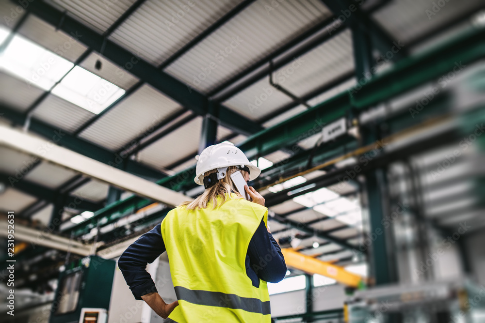 A rear view of an industrial woman engineer with smartphone in a factory, working. A rear view of an industrial woman engineer with smartphone in a factory, working.