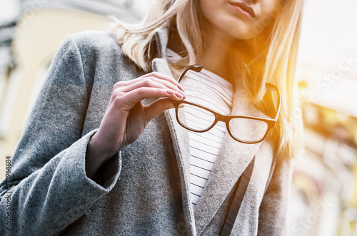 Closeup of stylish young woman holding eyeglasses in hand while standing on city street. Beautiful blonde hipster girl outdoors with spectaculars in her hand. Fashion eyewear for eye care