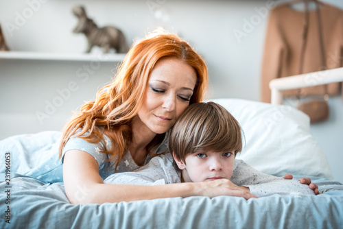 Young mother with her 2 years old little son dressed in pajamas are relaxing and playing in the bed at the weekend together, lazy morning, warm and cozy scene. Pastel colors, selective focus. photo