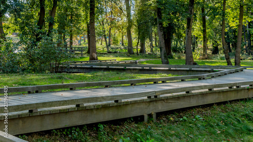 Zigzag wooden walkway surrounded by grass against green leafy trees in background, light sunlight between the foliage, park on a sunny summer day in Voerendaal, South Limburg, Netherlands photo