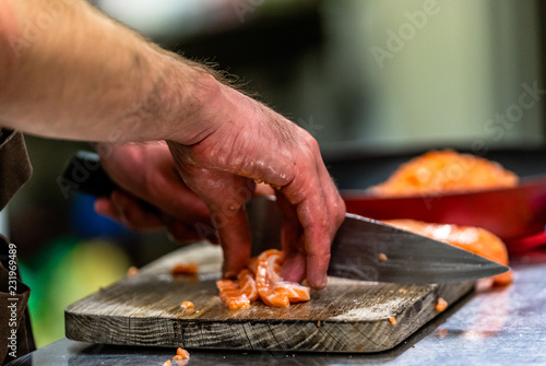 Male Chef Cutting Fresh Salmon on the Wooden Board with Blurred Pan in a Background