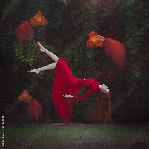 A beautiful girl with long red hair in a red dress is levitating above the ground. Surreal magic photo of a woman with big goldfish.
