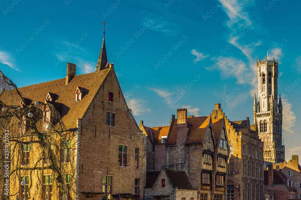 Medieval buildings with brick walls, gabled roof and Belfry bell tower in background against a blue sky, sunny day for tourism in Bruges, province of West Flanders, Belgium
