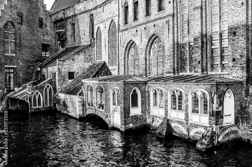 Medieval buildings on bank of the Dijver canal, brick walls, huge windows and enclosed terraces, calm water on a sightseeing day in province of West-Vlaanderen, Belgium. Black and white photography