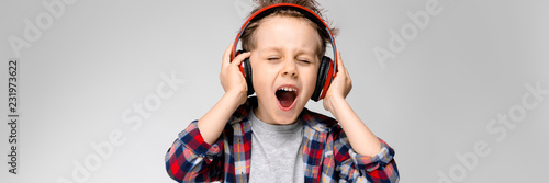 A handsome boy in a plaid shirt, gray shirt and jeans stands on a gray background. A boy in red headphones sings a song.