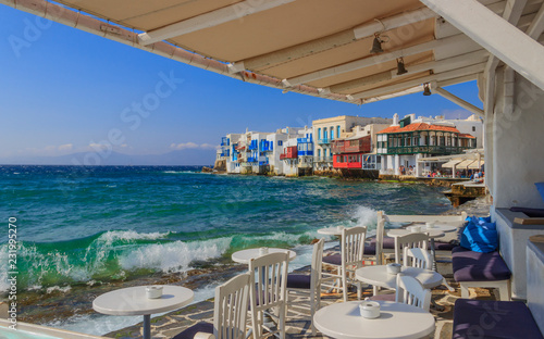 View of the famous pictorial Little Venice in Mykonos island. Splashing waves over bars and restaurants of old town, Cyclades, Greece. Chairs with tables in typical Greek restaurant.