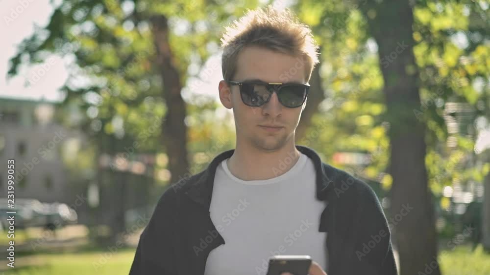 Front view of young man in sunglasses walking in the street, taking his smartphone from the pocket and starting to text. Tracking slow motion medium shot Front view of young man in sunglasses walking in the street, taking his smartphone from the pocket and starting to text. Tracking slow motion medium shot
