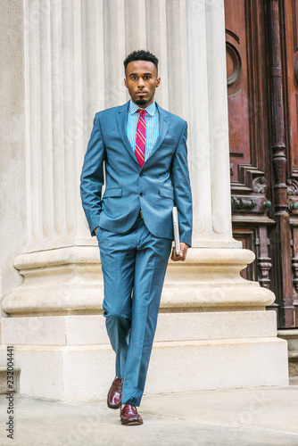 Obraz na płótnie Young African American Businessman with beard working in New York, wearing sky blue suit, violet red patterned tie, dark purple leather shoes, holding laptop computer, walking out from office door way w salonie