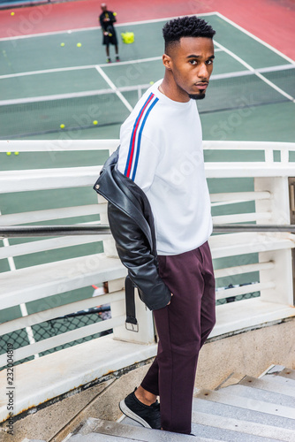 Man Casual Fashion in New York. Young Black Guy with beard, short afro hair, wearing white sport sweater, leather jacket off around arms, blank pants, standing on stairs with railing by tennis court..