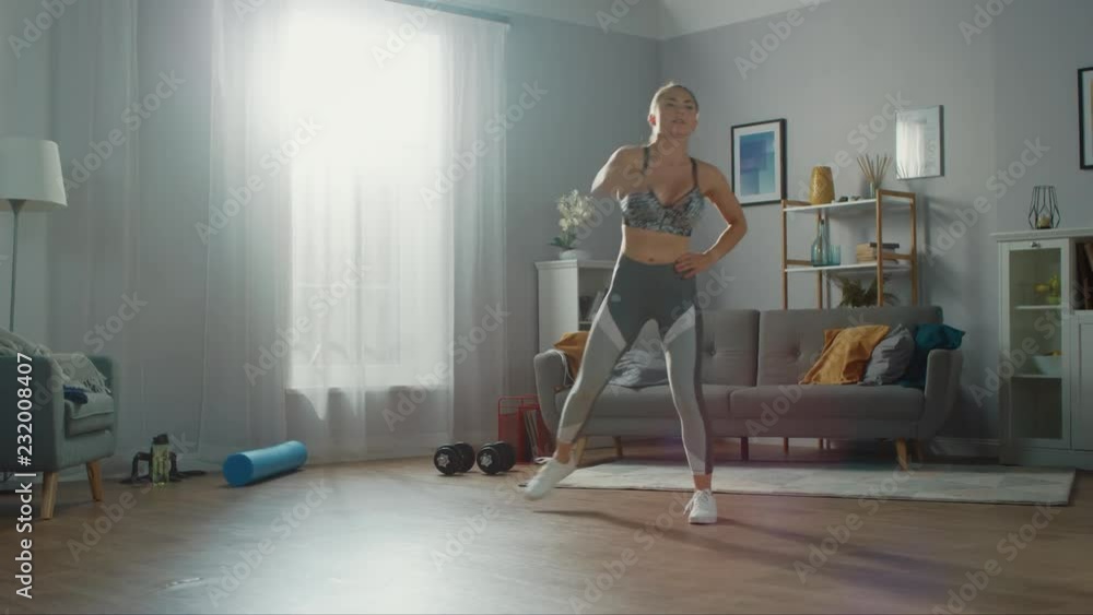 Shapely and Beautiful Athletic Fitness Girl in Sportswear is Doing Cardio Exercises in Her Bright and Spacious Living Room with Minimalistic Interior. Shapely and Beautiful Athletic Fitness Girl in Sportswear is Doing Cardio Exercises in Her Bright and Spacious Living Room with Minimalistic Interior.