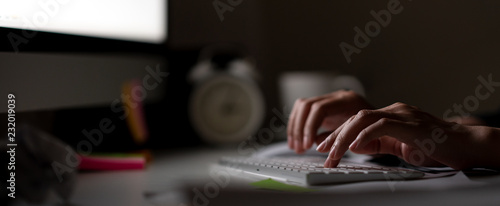 Workaholic hand typing keyboard in dark late night with computer screen light, clock, coffee cup and stuff on desk table with noir working in workplace or hacker cyber crime concept. Work hard banner.