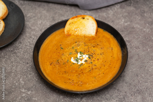 Cream soup of carrots and pumpkin with herbs, in a black bowl on a gray background.