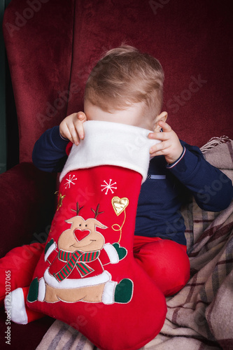 Cute lovely small boy holding a red christmas stocking sitting on the classic velvet red armchair