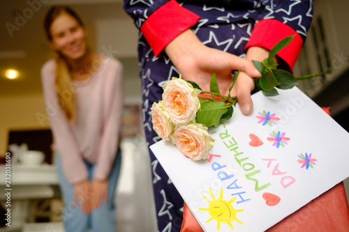 Child congratulates mother on Mother's Day, hides behind a gift with a postcard and flowers.