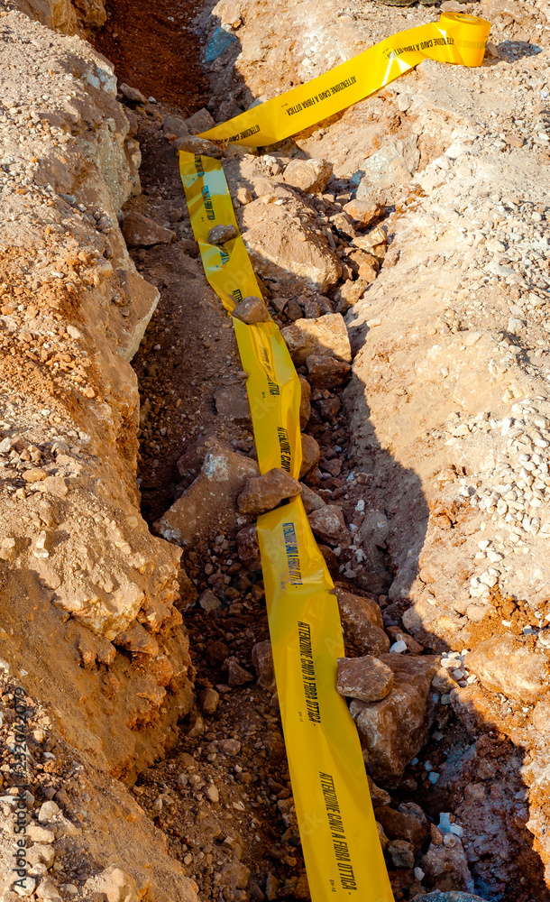 excavation with a narrow section to bury corrugated plastic cable ducts for fiber optic network..Yellow ribbon with the inscription excavation with a narrow section to bury corrugated plastic cable ducts for fiber optic network..Yellow ribbon with the inscription