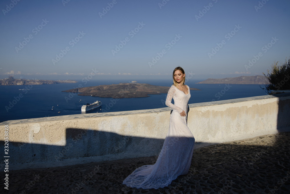 beautiful slim blond girl woman top model bride with long hair and legs in the white bridal ball gown with a long train and flounces stands on a picturesque island of Santorini in Greece