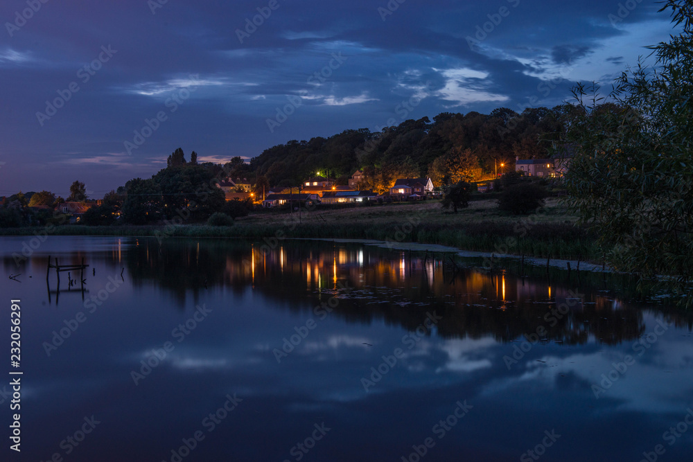 A Well Lit Village Viewed Across Water Against An Evening Sky, Fairburn, West Yorkshire