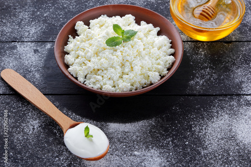 Cottage cheese in clay bowl and honey on wooden boards. Sour cream in wooden spoon. Dairy products and honey on dark background. Fresh mint and soft cheese for breakfast