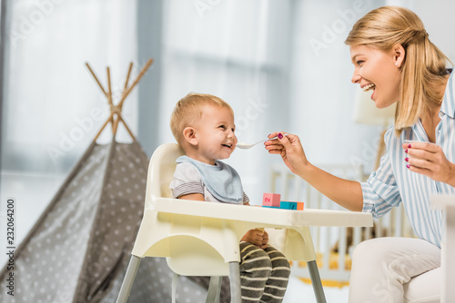 cheerful mom feeding son in highchair with baby food in nursery room photo