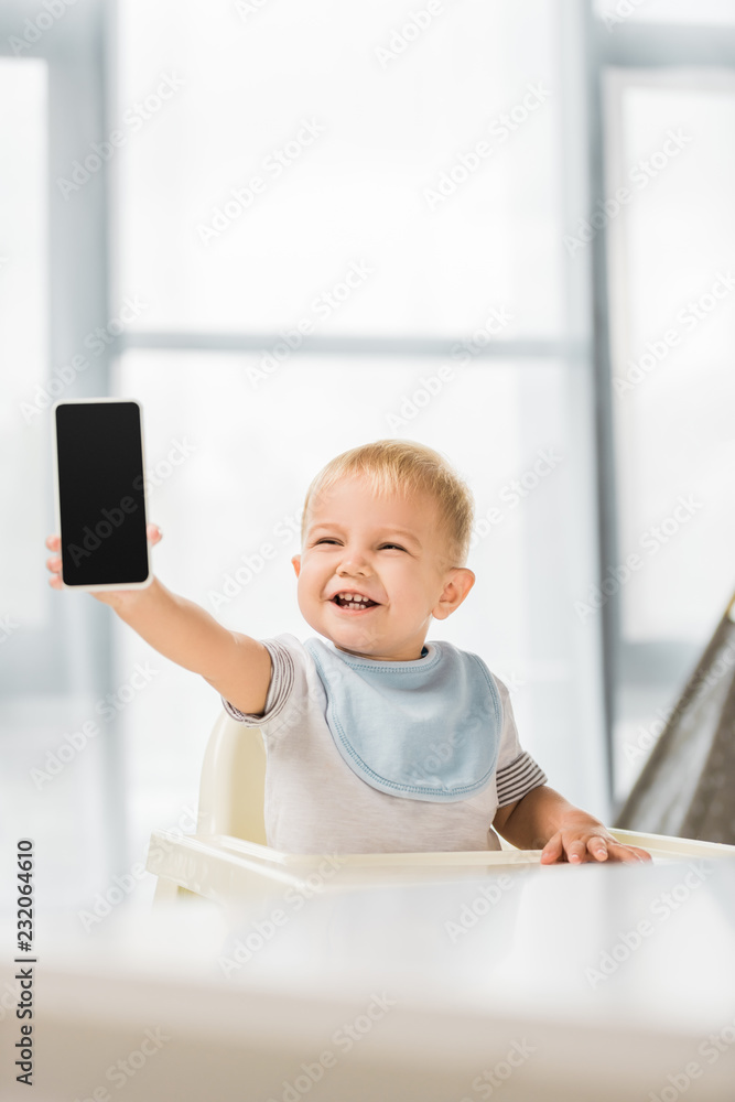 cute smiling toddler holding smartphone in hand and sitting in baby chair cute smiling toddler holding smartphone in hand and sitting in baby chair