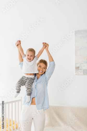 cheerful mother looking at camera and holding smiling toddler in hands in nursery room