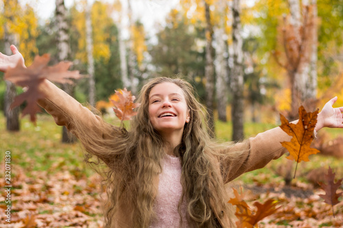 A teenager girl in a coat laughs and throws up leaves in autumn park against the background of yellow foliage.