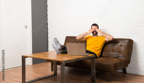 Man with his laptop in a room covering eyes by hands. Surprised to see what is ahead