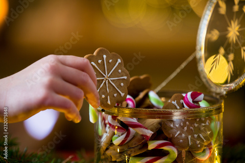 Cute little girl taking candies and gingerbread from glass jar and eating. Happy family holiday. Close-up hand. Living room decorated with lights and candles and Christmas tree. Holiday mood