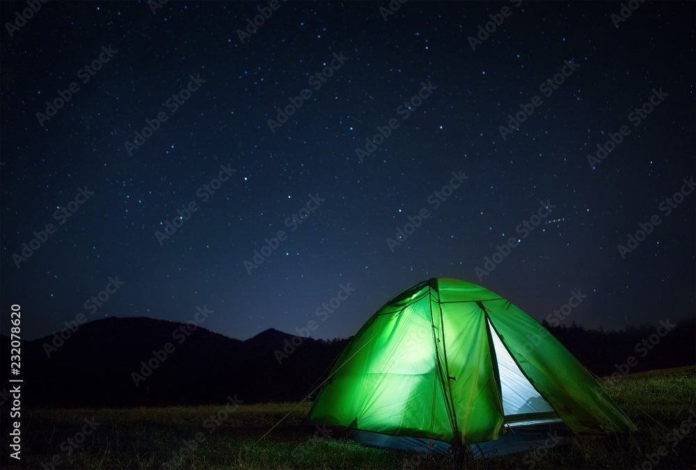 Camping tent with light inside is on the mountain valley under night starry sky