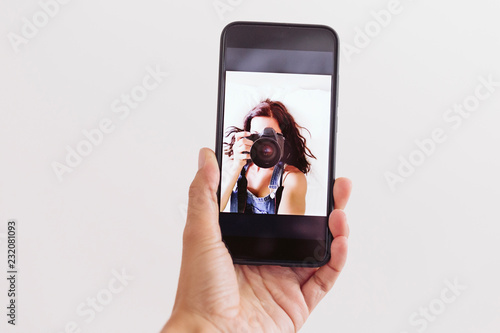 young woman taking a self portrait in front camera of the mobile phone at home. White walls and background. Reflex camera covering her face. Lifestyle