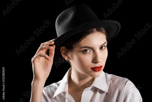 portrait of stylish woman in black hat and white shirt posing isolated on black