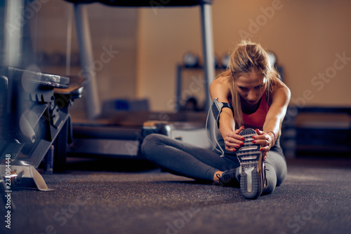 Fototapeta Naklejka Na Ścianę i Meble -  Woman stretching legs while sitting in a gym floor. In background exercise machines. Healthy lifestyle concept.