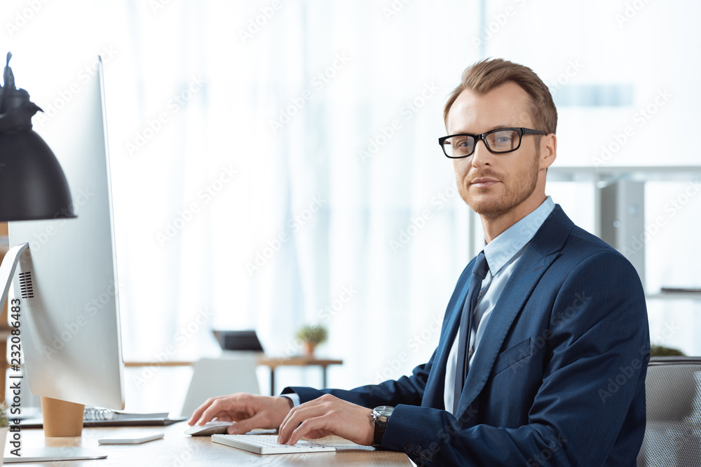 confident businessman in eyeglasses looking at camera and working at table with computer monitor in modern office