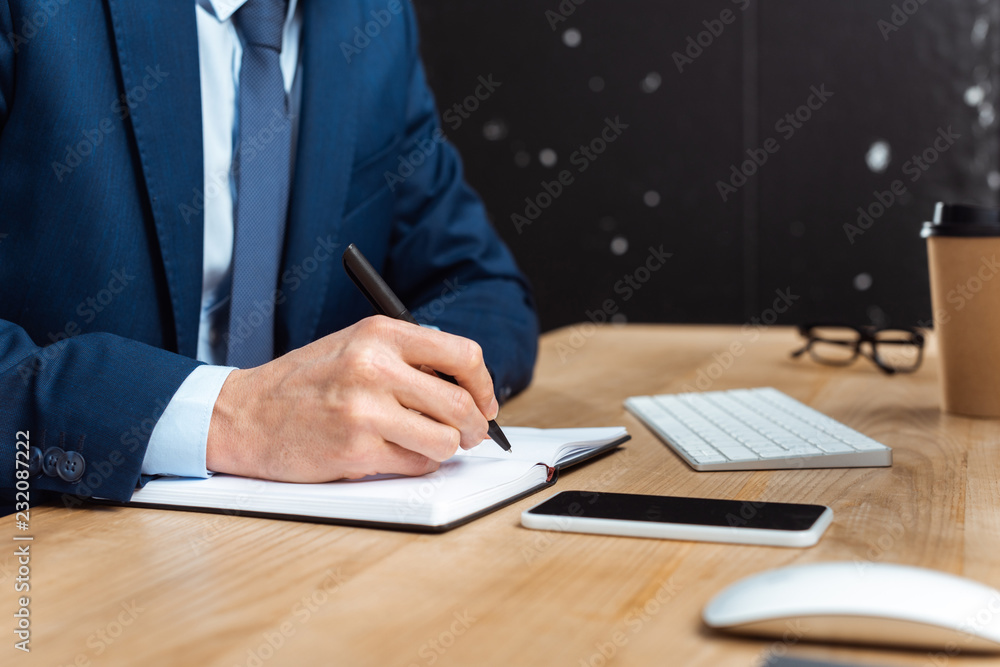 cropped image of businessman writing in textbook at table with smartphone in modern office cropped image of businessman writing in textbook at table with smartphone in modern office