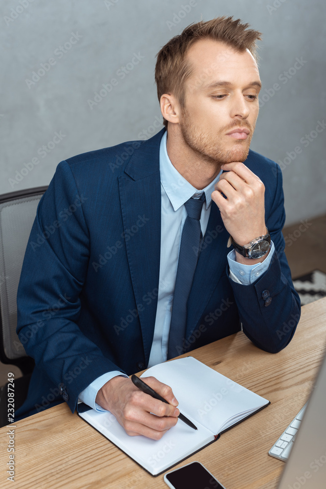 high angle view of thoughtful businessman writing in textbook at table with smartphone in modern office high angle view of thoughtful businessman writing in textbook at table with smartphone in modern office