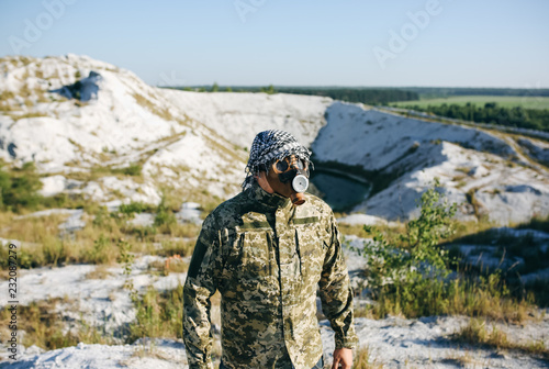 Soldier is standing in camouflage uniform and checkered keffiyeh shemagh bandana. Man in breathing mask safety respirator is outdoors in the abandoned deserted place. Environmental pollution concept.
