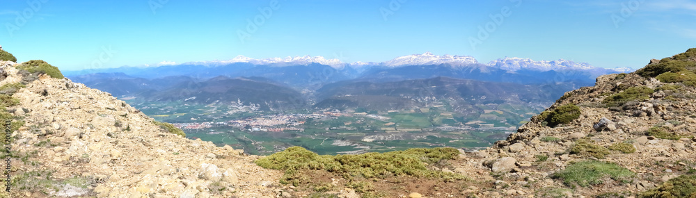 A wide angle landscape of snow-clad Pyrenees mountains and a wide valley with blue cloudy sky and some bushes in Peña Oroel, Aragon region, Spain