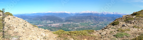 A wide angle landscape of snow-clad Pyrenees mountains and a wide valley with blue cloudy sky and some bushes in Peña Oroel, Aragon region, Spain