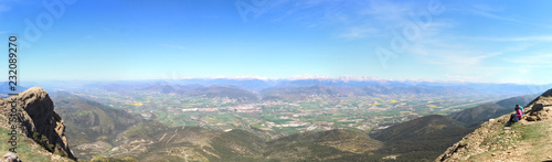 A wide angle landscape of snow-clad Pyrenees mountains and a wide valley with blue cloudy sky and some bushes in Peña Oroel, Aragon region, Spain