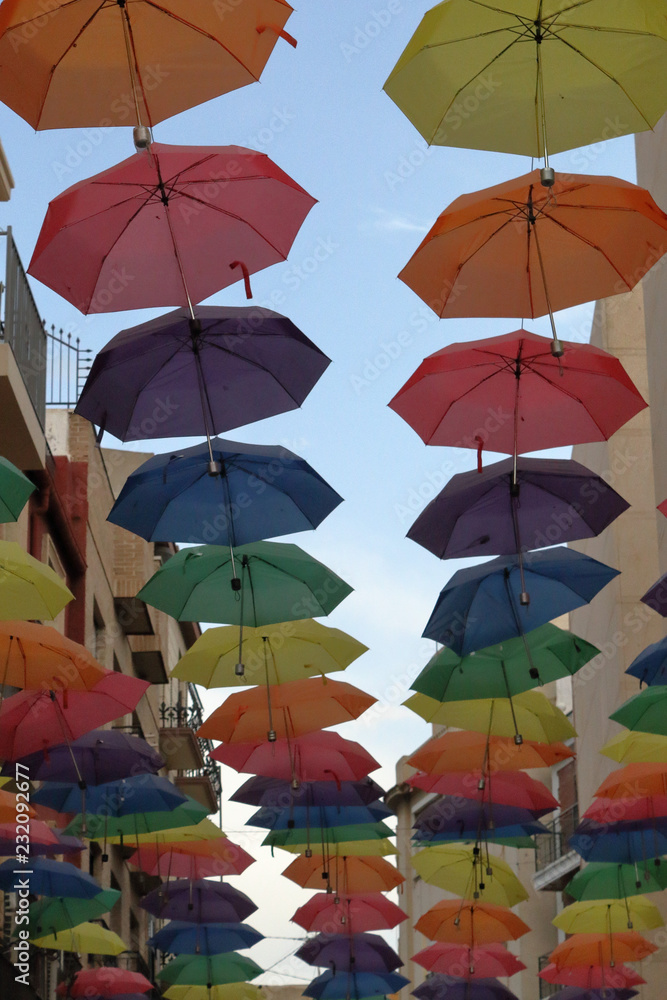 The sun shelter of an open air street through a series of coloured opened umbrellas hanged on cables, with a blue cloudy sky, in Orihuela, Spain