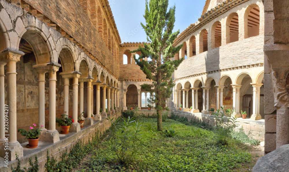 The cloister with romanesque arches, a fir tree and medieval frescos during a sunny day in the Colegiata de Santa Maria la Mayor of Alquezar, Spain