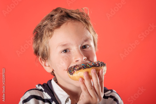 close up of one young blond and cute boy eating and holding with his hand one chocolate doughnut over a red background in studio. He has chocolate stains on his face.