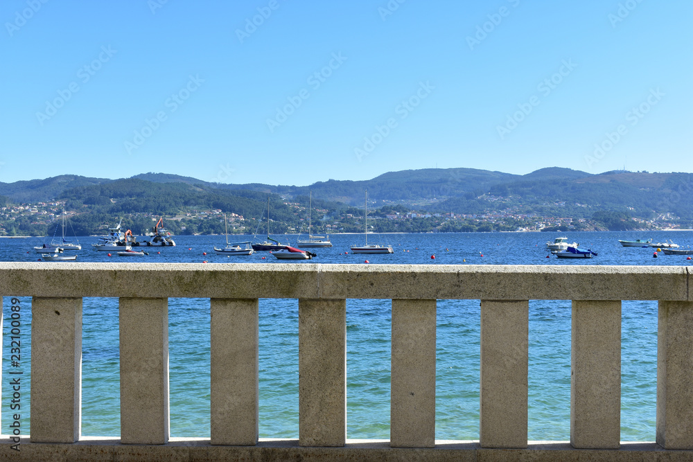 Stone handrail on a beach promenade. Turquoise water, boats and blue sky. Galicia, Rias Baixas, Spain.